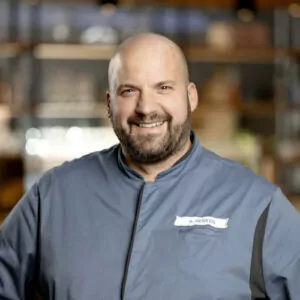 Ein l&auml;chelnder Koch in blauer Uniform mit Namensschild steht vor einem unscharfen Hintergrund im ATLANTIC Hotel.