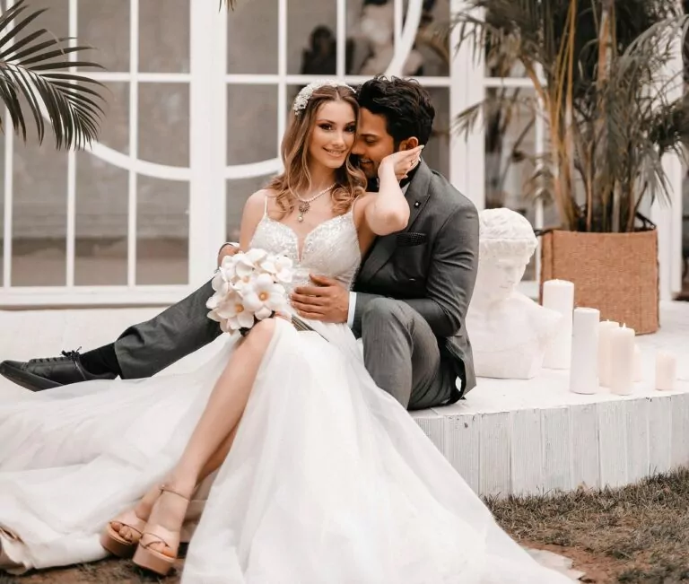 Ein Brautpaar sitzt gl&uuml;cklich vor einem eleganten Gew&auml;chshaus, umgeben von Pflanzen und Kerzen.