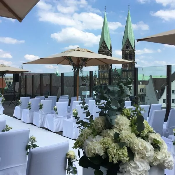 Dachterrasse mit Hochzeitssitzen, wei&szlig;en Blumen und Blick auf Bremer Domt&uuml;rme unter blauem Himmel.