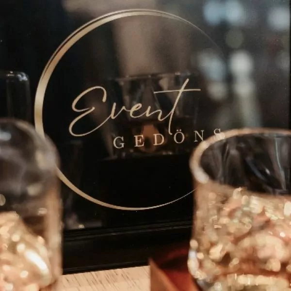 Nahaufnahme des Logos von "Event Ged&ouml;ns" im Bilderrahmen, im Vordergrund eine Kristall-Karaffe und Glas