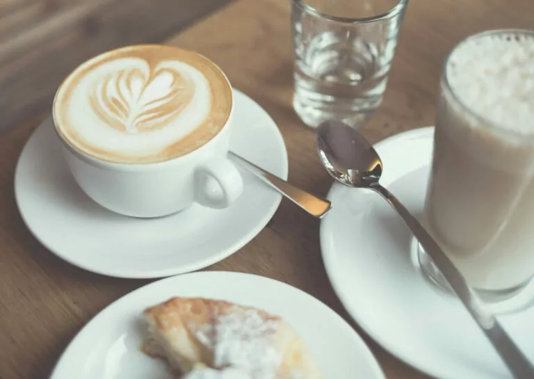 Tasse Cappuccino mit Latte Art, Glas Wasser, Latte Macchiato und Geb&auml;ck auf Holztisch.